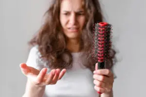 A woman holding a hairbrush with visible hair fall and frizzy strands in her hand, representing the comparison of Batana Oil vs Rosemary Oil for treating frizzy, damaged hair and improving hair strength and scalp health.